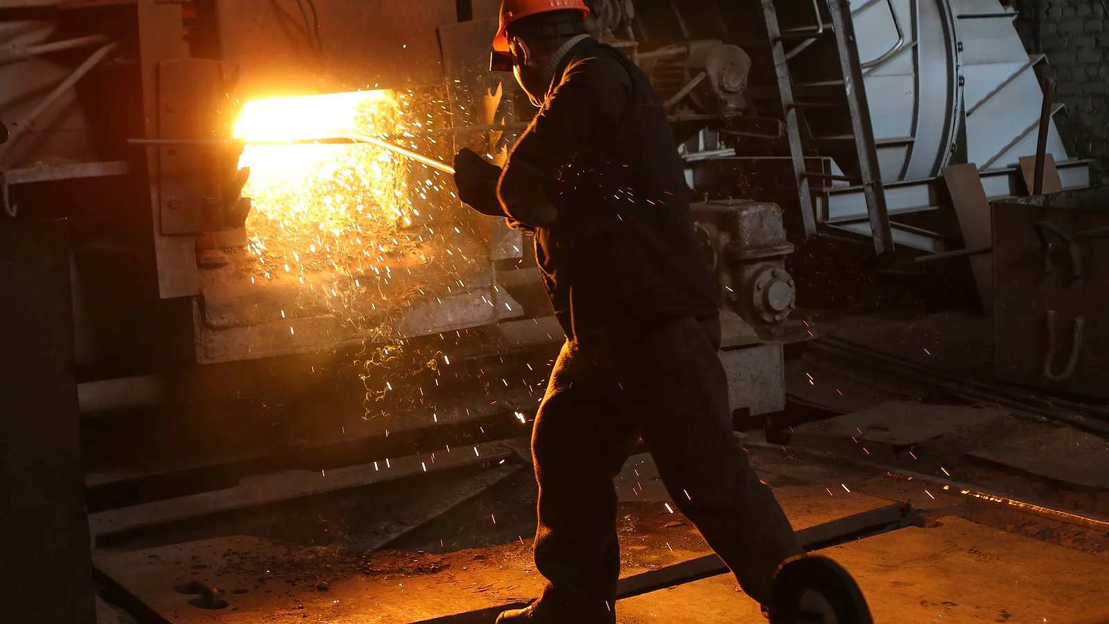 Steel plant worker handling intensely heated metal beside industrial production equipment